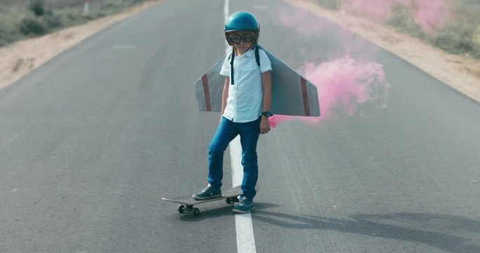 CINEMAGRAPH - seamless loop. Little boy wearing helmet and styrofoam wings standing on a skateboard on a rural road, pretending to be a pilot