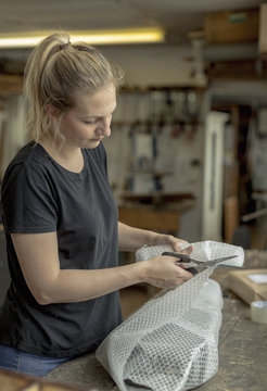 Blond woman standing in a workshop, cutting bubble wrap with pair of scissors, wrapping a parcel for delivery or posting. 