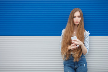 Emotional portrait of a adult pretty blonde woman with gorgeous extra long hair posing outdoors against blue grey striped metal background wearing blue blouse high waist jeans holding smartphone