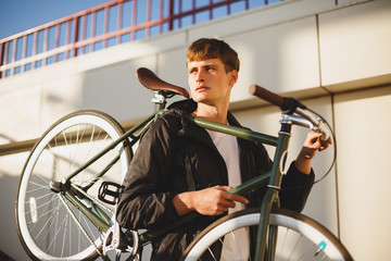 Portrait of thoughtful boy with brown hair standing and holding bicycle while dreamily looking aside. Young man in down jacket standing with classic beautiful bicycle on street