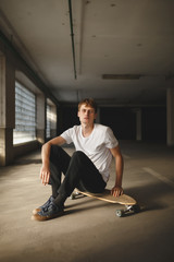 Fototapeta na wymiar Portrait of cool boy with brown hair sitting on skateboard and dreamily looking in camera. Young thoughtful man in white t-shirt sitting and holding cigarette in hand while posing on camera