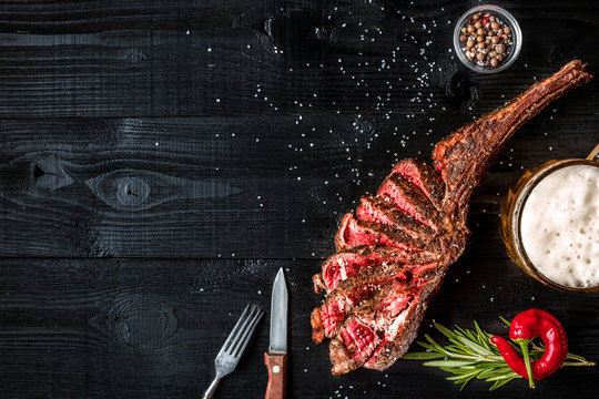 Barbecue dry aged rib of beef with spice, vegetables and a glass of light beer close-up on black wooden background