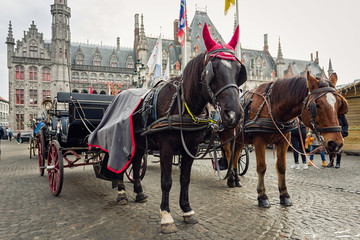 Obraz premium Horse-drawn carriage waiting on tourists for a guided tour on the center square in Bruges, Belgium. Christmas theme hat and ear covers on the horse, Christmas market on the background