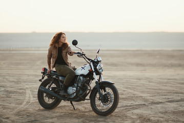  young beautiful woman sitting on her old cafe racer motorcycle in desert at sunset or sunrise