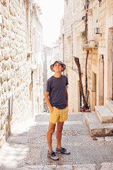 Handsome attractive young man in yellow shorts and panama hat, walks around old european town in summer heat, explores ancient tourist attractions