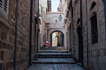 Fototapeta na wymiar Narrow steep street of beautiful ancient european village or city, Dubrovnik Croatia with cobbled paved walls and floors. Lonely lost tourist sits on steps, exploring