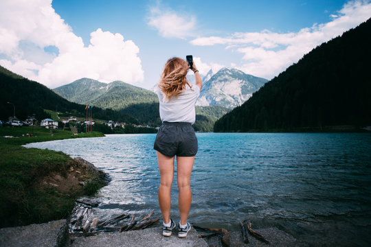 Young female tourist millennial adventurer stands on shore of beautiful pristine lake high in mountains with turquoise water, she makes photo on her smartphone for memories of travel