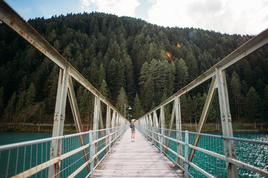 Young fit adventurer man with camera makes photo of nature, stands in middle of metal pedestrian bridge over pristine blue water lake that connects two shores, forest and city