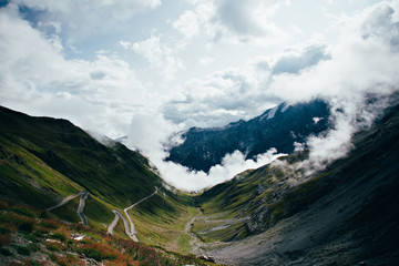 Shot from top of Stelvio pass between Italy and Switzerland on sunny day, standing above clouds and overlooking hairpins and sharp turns that lead to summit