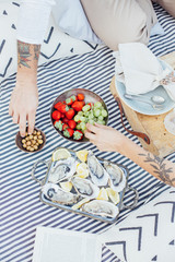 Unconventional hipster couple enjoys luxurious snack of fresh oysters during romantic date on picnic blanket, in chic and dreamy setting with fruits and olives