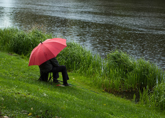  Senior man is sitting  under a red umbrella and fishing  on the lake bank on a autumn rainy day.