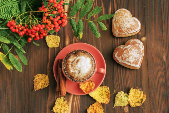 a cup of coffee cappuccino with cinnamon, gingerbread in heart-shaped form, yellow leafy leaves on a wooden table