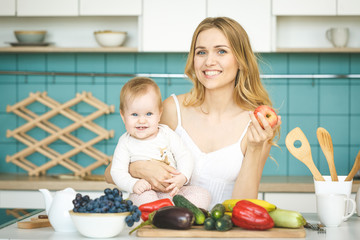 Young mother looking at camera and smiling, cooking and playing with her baby daughter in a modern kitchen setting. Healthy food concept.