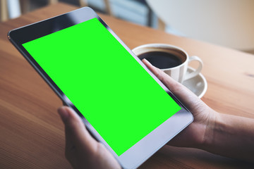 Mockup image of business woman's hands holding black tablet pc with blank green screen and coffee cup on wooden table in cafe background