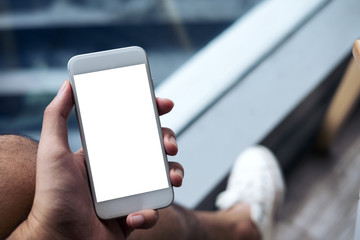 Mockup image of a man's hand holding white mobile phone with blank screen on thigh with white canvas shoes in modern cafe with feeling relax