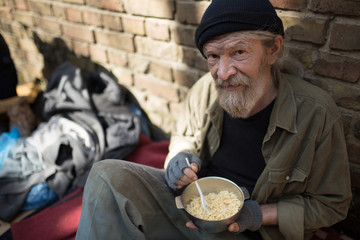 Man with no home, sitting by the brick wall. Homeless man with metal bowl with meal.