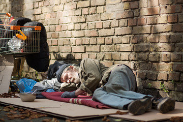 Sleeping homeless man lying on cardboard. Tramp lying by the brick wall, his posessions on the...