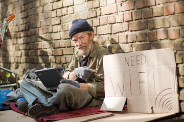 Homeless man pointing on cardboard with sign need wi fi. Tramp sitting on cardboard by the brick...