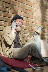 Side view of a homeless man sitiing by the brick wall, talking on phone. Tramp holding cell phone in one hand and solar battery in another.