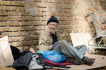 Tramp sitting in the street, his thigns scattered around him. Old homeless man sitting on cardboard.