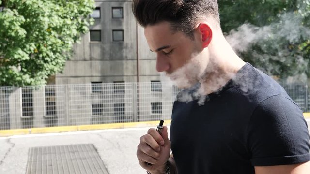 Handsome man standing in city street and vaping with electronic cigarette.