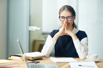 Waist-up portrait of thoughtful young white collar worker in eyeglasses sitting at office desk and thinking over start-up project, blurred background