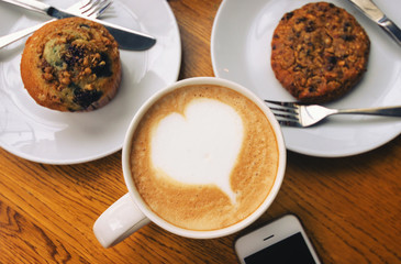 coffee with heart love shape in white cup with chocolate cupcake and smartphone on wood table.