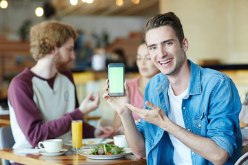 Fototapeta na wymiar Handsome young man with wide smile posing for photography while pointing at blank screen of modern smartphone, his friends chatting animatedly with each other and enjoying lunch at cozy cafe