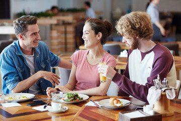 Multi-ethnic group of cheerful friends gathered together at lunchtime in cozy small cafe and chatting animatedly with each other, blurred background