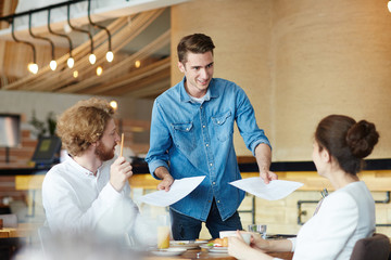 Portrait of smiling young manager wearing denim shirt passing documents to his colleagues while working together at cozy small cafe