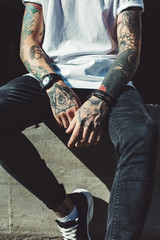 Faceless shot of young man with tattooed hands posing on concrete fence outside in sunlight.