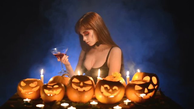 Sexy woman in Halloween costume and makeup sitting at table with pumpkins over dark blue background playing with cocktail