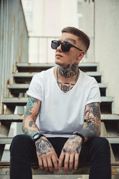 Confident tattooed man in white t-shirt and sunglasses sitting on stairs outside looking away.