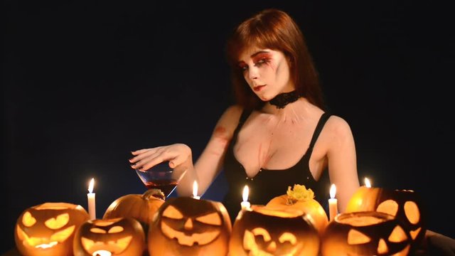 Sexy woman in Halloween costume and makeup sitting at table with pumpkins over dark blue background playing with cocktail