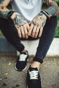 Faceless shot of young man with tattooed hands posing on concrete fence outside in sunlight.