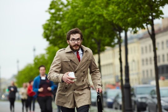 Busy man in elegant trench-coat carrying drink and briefcase while running for work after coffee-break