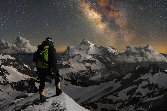 Night photo climber stands on top of a mountain in the snow and looks at the surrounding mountains over which the starry sky and the milky way