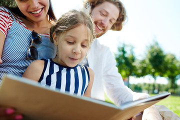 Adorable girl and her parents reading interesting book together in park