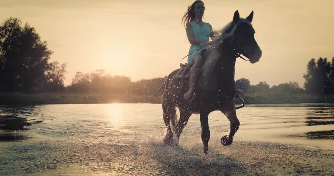 Slow motion shot. Woman riding horse on the background of beautiful sunset and water. Girl model on horseback on river water at sunset, backlit with sunshine.