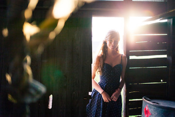 Young girl leaning against doorway of a rustic barn with the sunset behind her