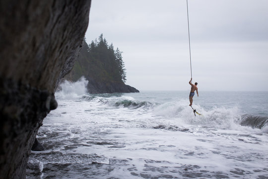 Man hanging on a ropeswing over crashing ocean waves