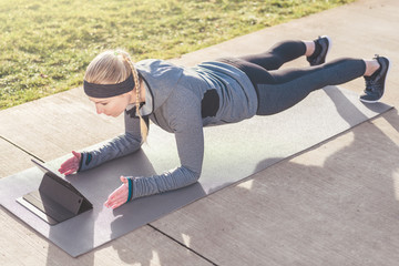 Naklejka premium High-angle view of a fit young woman watching a motivational video on tablet PC, while exercising the forearm plank position on a mat outdoors in the park