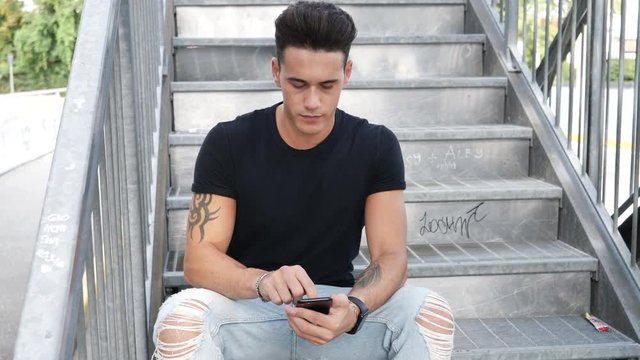 Handsome young man standing outdoors in urban environment on metal stairs, calling on cell phone