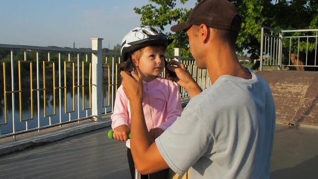A man is putting a helmet on a child. The father wears a helmet on his daughter's head.
