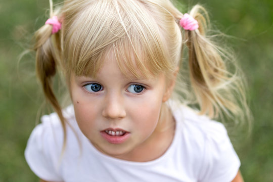 Close Up Portrait Of Cute Adorable Little Blonde Caucasian Girl In A White T-shirt In A Park, Looking Aside, Thinking Or Dreaming Of Something, Puzzled Or Astonished, Happy Childhood Concept.