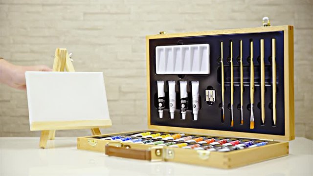 Person hand puts mini easel with canvas beside suitcase with paint tubes 4K