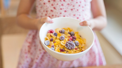 Morning breakfast, little girl hand holding cornflakes cereal, strawberry, blackberry and milk in a white bowl