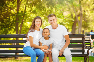 Young family with adopted African American boy sitting on bench in park