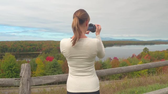 Young brunette traveler taking photos of breathtaking nature in autumn. Girl tourist photographing stunning lake surrounded by colorful autumn forest. Female explorer on leaf-peeping trip on viewpoint