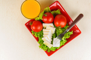 Tomatoes and cheese slices lie on a sheet of fresh salad in a red plate. A glass of orange juice stands side by side on the table.